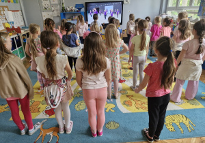 Dziewczynki stoją przed monitorem multimedialnym.