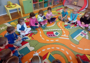 Dzieci siedzą na dywanie w kole i podają sobie piłkę.
