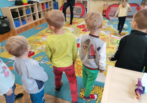 Dzieci stoją na dywanie. Dziewczynka kopie piłką do bramki.