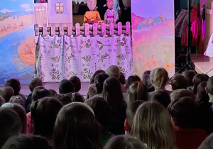 Na scenie mężczyzna z dwoma lalkami. Dzieci oglądają przedstawienie teatralne.