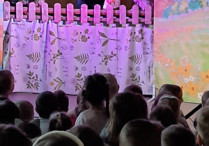 Na scenie mężczyzna z lalkami przedstawiającymi Anulkę i Kózkę. Dzieci oglądają przedstawienie teatralne.