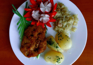 Na stoliku leży talerz, na którym znajdują się: ziemniaki, kotlet schabowy, gotowana kapusta, rzodkiewka i papryka.