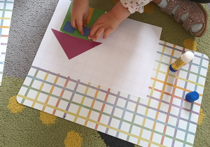 Dziewczynka przykleja okno do swojego domku z figur geometrycznych.