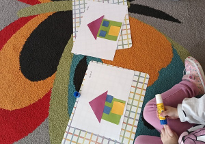 Dzieci siedzą na dywanie, przed nimi leżą kartki z przyklejonymi domkami z figur geometrycznych.