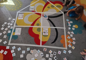 Dzieci siedzą na dywanie, na środku koła widać domek wyklejony z taśmy malarskiej, podzielony na pomieszczenia oraz obrazki różnych przedmiotów.