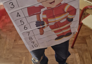 Chłopiec prezentuje wykonaną przez siebie pracę - strażaka pociętego na części, ułożonego i przyklejonego na kartkę.