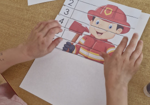 Dzieci przy stoliku przyklejają na kartkę puzzle przedstawiające strażaka.