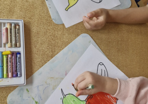 Dzieci kolorują pastelami kontury owoców w koszyku.