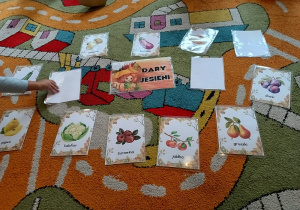 Na dywanie leżą obrazki warzyw i owoców, dziecko odsłania kolejny obrazek.