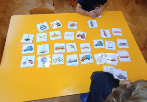 Dzieci siedzą przy stoliku, na których leżą obrazki z różnymi środkami transportu.