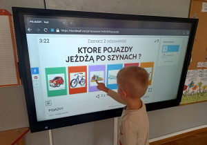 Chłopiec wykonuje zadanie na tablicy multimedialnej – wskazuje pojazd poruszający się po szynach.