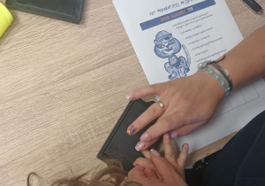 Dziecko z pomocą pani policjantki odbija swoje paluszki w tuszu, by wykonać odciski palców.
