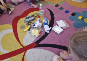 Dzieci siedzą w kole. Na środku rozrzucone różne odpady oraz klocki w kolorach koszy do segregowania odpadów.