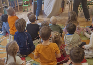 Dzieci siedzą na dywanie. Kobiety wyciągają wielką maskotkę - stonogę, z worka.