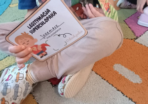 Chłopiec siedzi na dywanie, trzymając w ręce "Legitymację superchłopaka".