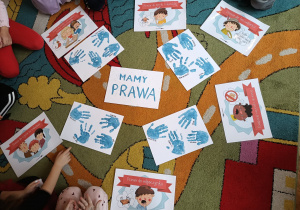 Na dywanie rozłożone ilustracje przedstawiające różne prawa oraz odbite na kartkach dłonie dzieci.
