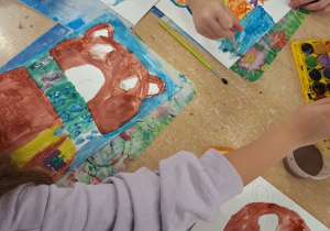 Dzieci siedzą przy stoliku. Każde dziecko maluje farbami akwarelowymi portret misia na białym kartonie.