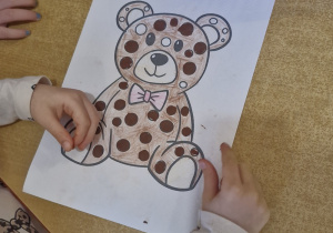 Dziewczynka pracuje przy stoliku - wykleja pokolorowanego na brązowo misia plasteliną w wyznaczonych miejscach.