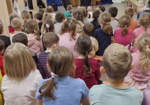 Dzieci siedzą na ławeczkach. Na scenie mężczyzna z mikrofonem.