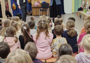 Dzieci siedzą na ławeczkach i obserwują jak kobieta gra na oboju.