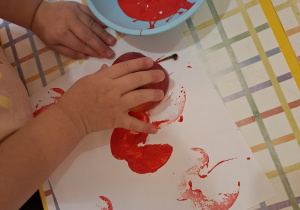 Dziecko stempluje jabłkiem zamoczonym w czerwonej farbie na kartce.