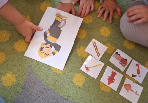 Na dywanie wokół dzieci ilustracja przedstawiająca zawód strażaka oraz jego atrybuty.