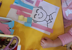 Dziewczynka przykleja na kartkę kolorowe włosy z papieru.