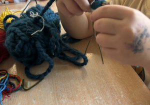 Dziecko owija patyki w kształcie ludzika niebieską włóczką.