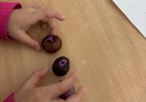 Dziecko przy stoliku trzyma kasztany, na których wypisane są samogłoski.