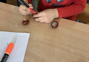 Dziecko przy stoliku pisze po kasztanach mazakiem.