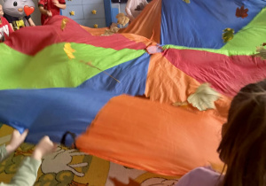 Dzieci trzymają chustę animacyjną i podrzucają na niej liście.