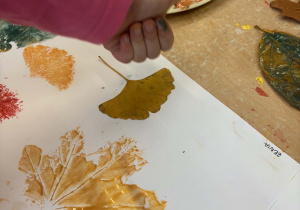Dzieci pracują przy stoliku - odbijają na białej kartce liście zamoczone w kolorowej farbie.