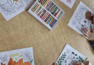 Dzieci przy stoliku kolorują pastelami wiewiórkę.