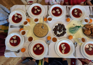 Dzieci siedzą przy świątecznym stole. Na talerzach mają nalany czerwony barszcz z uszkami.