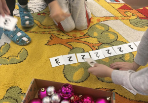 Dzieci na dywanie układają plakietki z cyferkami; w pudełku kolorowe bombki.