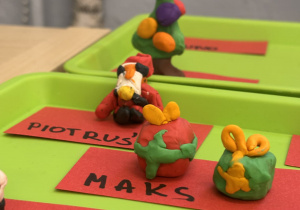 Wylepione z plasteliny figurki prezentów , Mikołaj oraz choinka na zielonych podkładkach z czerwonymi karteczkami z imionami dzieci.