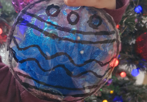 Dziecko pokazuje wykonaną przez siebie sesnoryczną bombkę z folii do laminowania, żelu i brokatu.