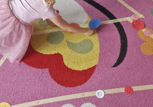 Dziecko układa papierowe bombki na choince wyklejonej taśmą malarską na dywanie.