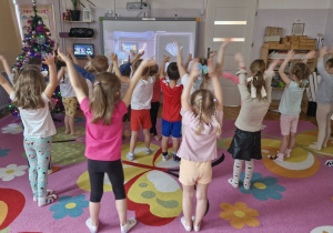 Dzieci z rękoma uniesionymi do góry przed tablicą multimedialną.
