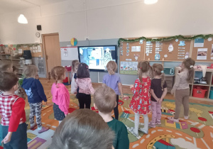 Dzieci z grupy zielonej ćwiczą przed tablicą multimedialną.