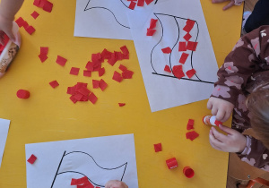 Dzieci klejem przyklejają czerwoną bibułę na kartkę papieru.