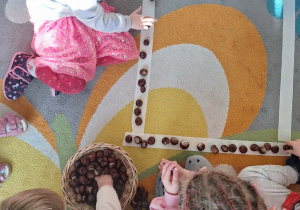 Dzieci układają na dywanie kwadrat z kasztanów.