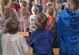 Dzieci powtarzają ruchy za prowadzącym, kobieta gra na instrumencie.