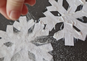Dziecko posypuje solą śnieżynki wycięte z papieru i przyklejone na kartkę.