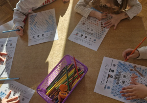 Dzieci przy stoliku wykonują kartę pracy - kolorują zakodowanego bałwanka.