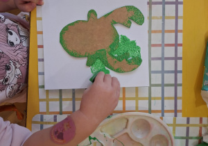 Dziewczynka stempluje gąbką na białej kartce papieru zieloną farbą.