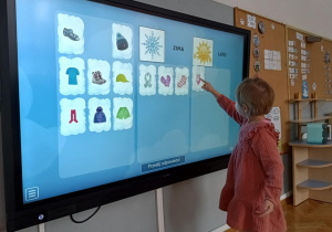 Dziewczynka na tablicy multimedialnej dopasowuje cechy charakterystyczne do podanych pór roku.