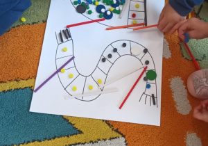 Dzieci w grupie na dywanie układają dary Froebla na ilustracjach przedstawiających czapkę i szalik.