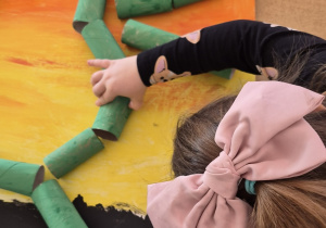 Dziewczynka przykleja na karton rolkę po papierze toaletowym pomalowaną na zielono.