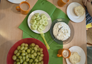 Dzieci siedzą przy stole - na stole bułki, winogrono, ogórki.